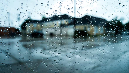 A close-up view of raindrops on a glass pane, with a blurred background of a house and wet pavement, creating a serene atmosphere in this HD photography wallpaper.
