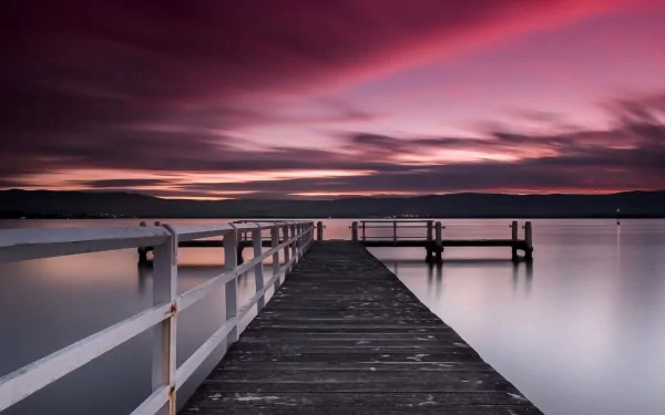 A serene pier extends into calm waters under a vibrant sunset, creating a tranquil scene. This HD wallpaper captures the beauty of man-made structures in nature.