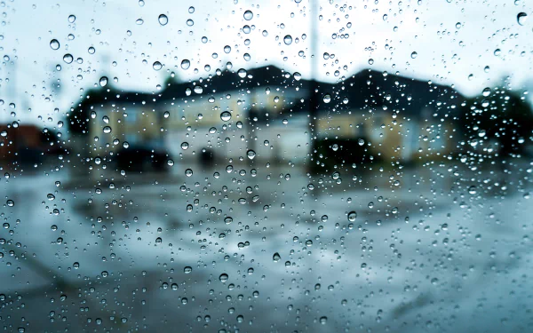 A close-up view of raindrops on a glass pane, with a blurred background of a house and wet pavement, creating a serene atmosphere in this HD photography wallpaper.
