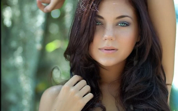 A high-definition desktop wallpaper featuring a woman with long, dark hair and blue eyes, posing outdoors with a serene and confident expression.