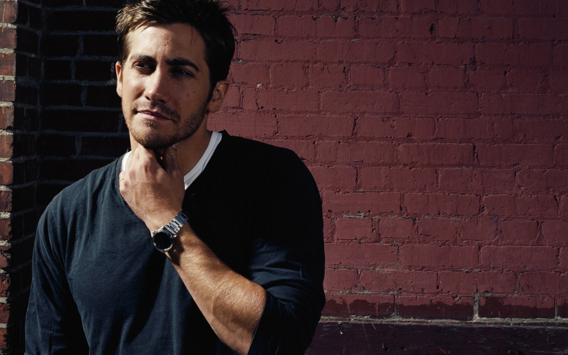 HD desktop wallpaper featuring celebrity Jake Gyllenhaal posing against a brick wall, wearing a dark shirt and watch, with a confident expression.