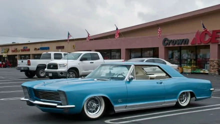 HD desktop wallpaper featuring a classic blue 1965 Buick Riviera parked in front of a strip mall with pickup trucks and storefronts in the background.