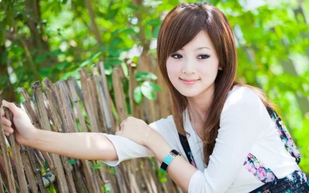 HD desktop wallpaper featuring Mikako Zhang Kaijie, a woman with brown hair in a white top, leaning against a wooden fence with a green leafy background.