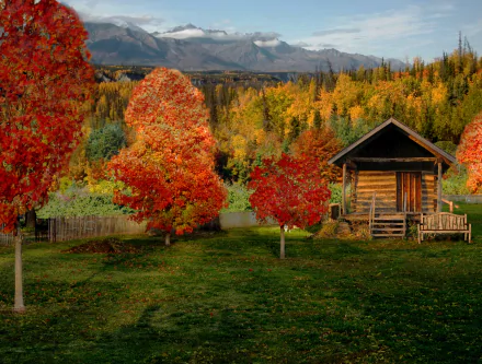 HD desktop wallpaper showing a man-made rustic cabin surrounded by vibrant autumn trees with colorful foliage against a backdrop of mountains and cloudy sky.