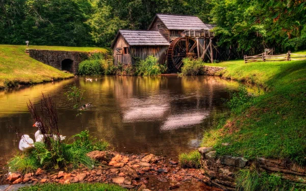 HD desktop wallpaper of a serene man-made watermill beside a calm pond, surrounded by lush greenery and a wooden fence.