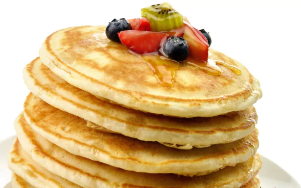 Close-up HD desktop wallpaper of a stack of fluffy pancakes topped with syrup, sliced strawberries, blueberries, and kiwi on a white background.