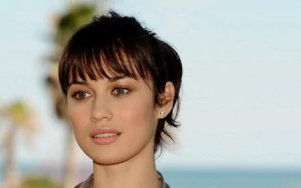 A stunning HD desktop wallpaper featuring celebrity Olga Kurylenko, with soft natural lighting and a blurred beach background, highlighting her expressive features.