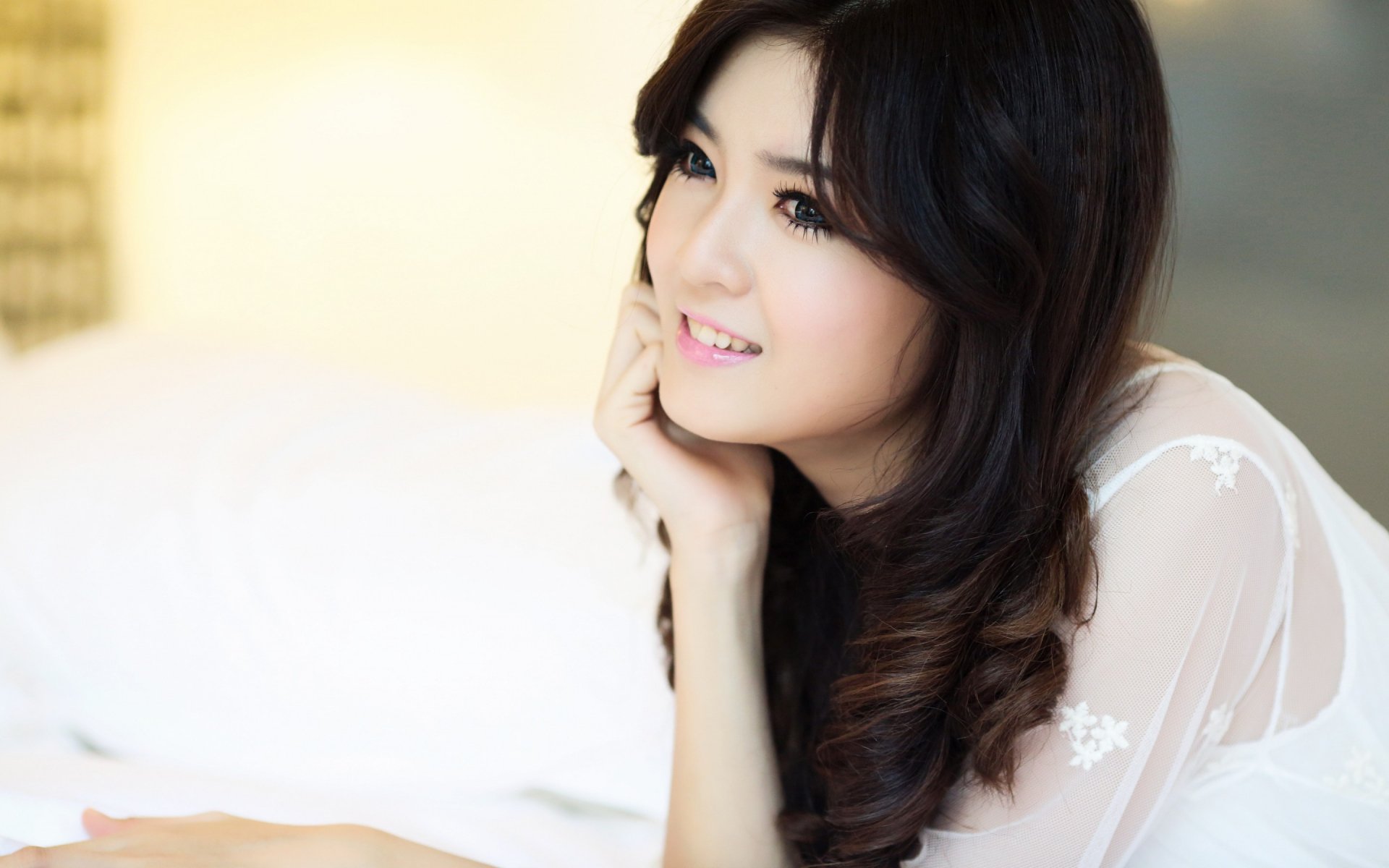 HD desktop wallpaper featuring a woman in white attire, smiling while resting her chin on her hand, in a softly lit setting.