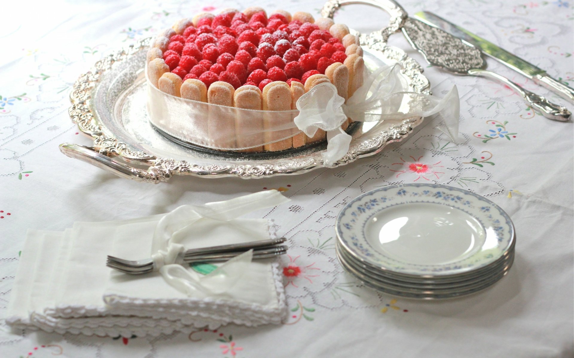 A beautifully arranged cake topped with fresh raspberries on a silver platter, accompanied by elegant plates and utensils, set against a floral tablecloth. A delightful food-focused desktop wallpaper.