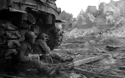 Two soldiers take cover beside a military vehicle, surrounded by the ruins of World War II. This striking black-and-white scene serves as a compelling HD desktop wallpaper.