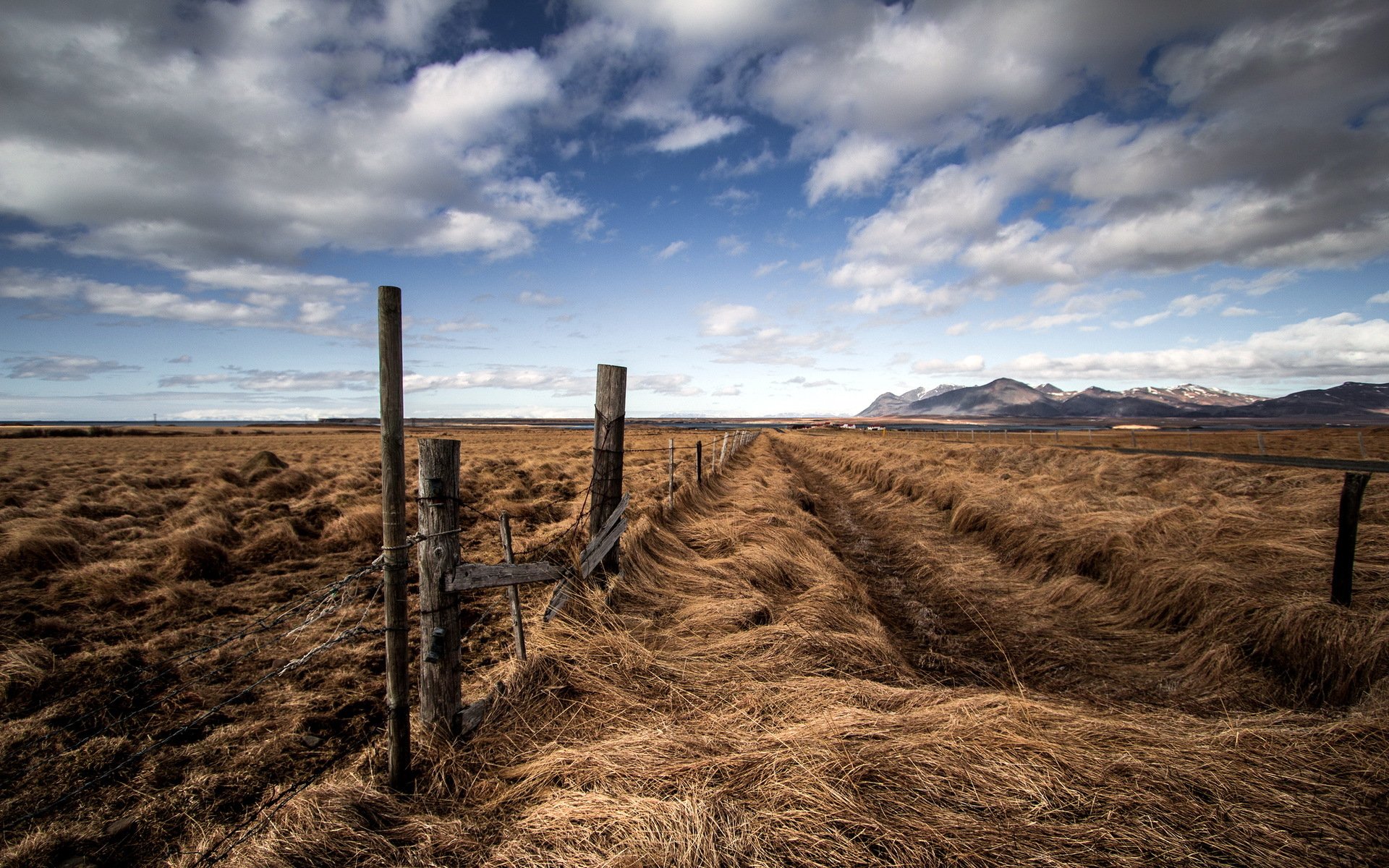 HD desktop wallpaper featuring a vast field with dry grass, weathered fence posts, and a dramatic cloudy sky in a natural landscape.