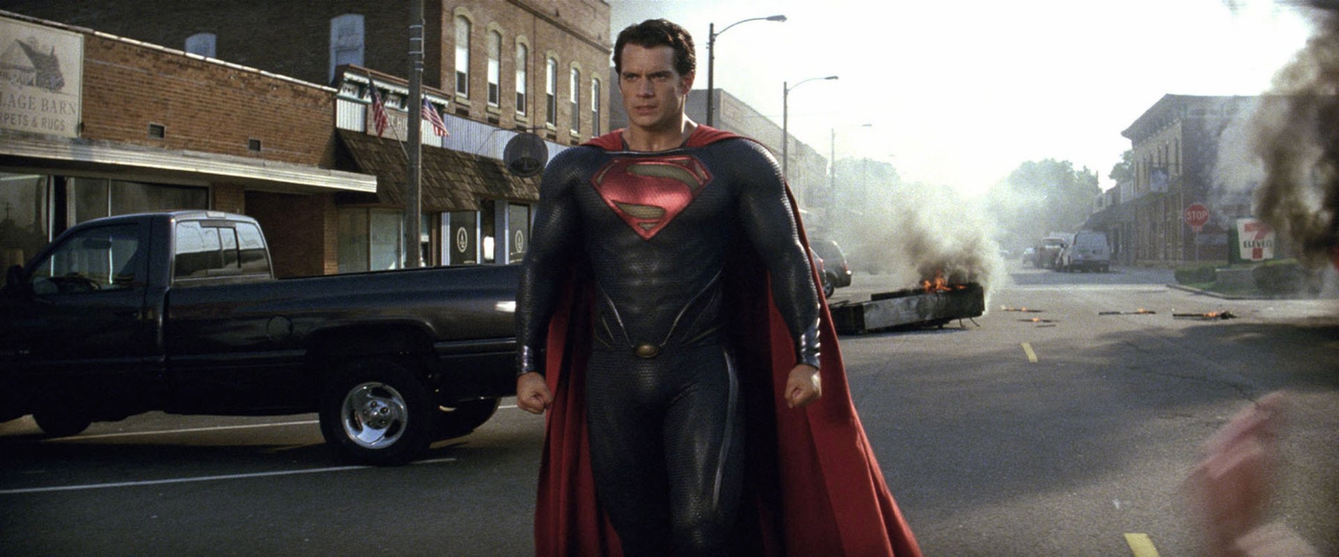 A dynamic HD wallpaper featuring Henry Cavill as Superman in Man of Steel, striding confidently through a street with a fiery explosion in the background.