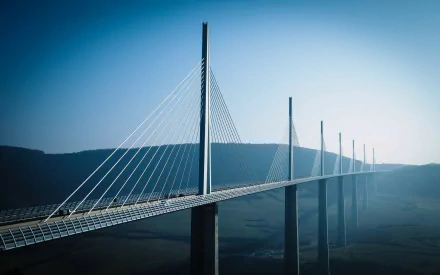 A breathtaking view of the Millau Viaduct, a man-made bridge, stretches elegantly across the landscape, framed by a serene blue sky. Ideal HD wallpaper for nature and engineering enthusiasts.