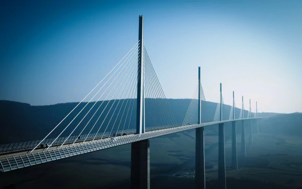 A breathtaking view of the Millau Viaduct, a man-made bridge, stretches elegantly across the landscape, framed by a serene blue sky. Ideal HD wallpaper for nature and engineering enthusiasts.