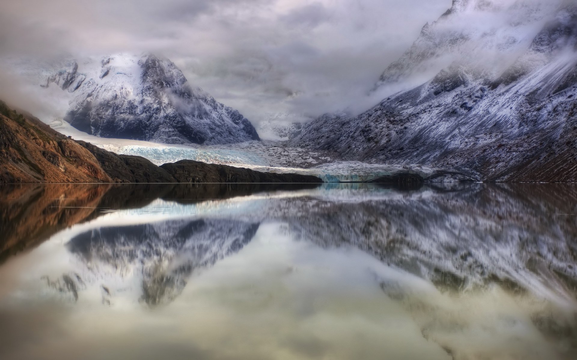 HD desktop wallpaper showcasing a serene lake in Argentina, surrounded by snow-capped mountains and dramatic cloud-covered skies reflecting on the water’s surface.