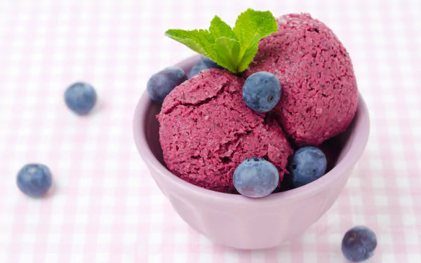A charming HD wallpaper featuring a bowl of purple ice cream garnished with mint leaves and surrounded by fresh blueberries, set against a pastel checkered background.