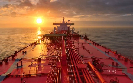 A breathtaking view of an oil tanker navigating tranquil waters at sunrise, with vibrant colors reflecting off the ship's deck against a dramatic sky backdrop.