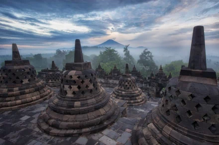 HD desktop wallpaper of the ancient Borobudur temple at sunrise, showcasing its religious stupas enveloped in mist with a dramatic sky and distant mountains.