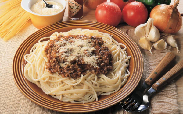A delicious plate of pasta topped with meat sauce and cheese, surrounded by fresh tomatoes, garlic, and onions, set against a rustic backdrop, serving as an enticing HD desktop wallpaper.