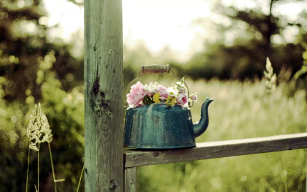 A vintage teapot filled with colorful flowers rests on a wooden fencepost, set against a lush, green landscape, creating a serene and charming atmosphere.