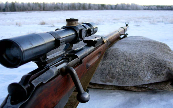 HD PC desktop wallpaper/background: man-made rifle with scope on a sandbag in a snowy field, barrel pointing toward a distant tree line.