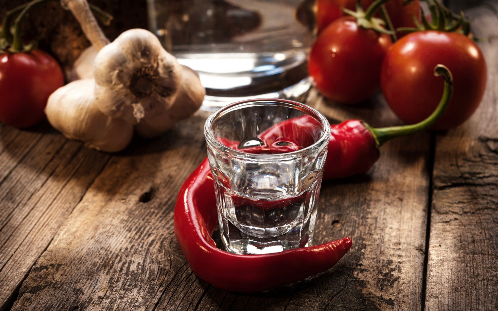 HD PC desktop wallpaper featuring a shot glass of clear drink surrounded by red chili peppers, garlic bulbs, and ripe tomatoes on a rustic wooden surface.