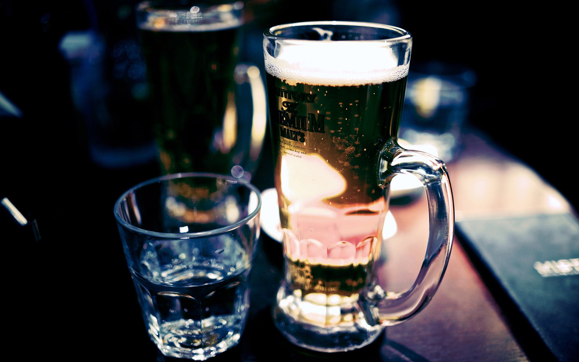 HD desktop wallpaper featuring a close-up of a frosty beer mug and a glass of water on a dark wooden table in a cozy setting.
