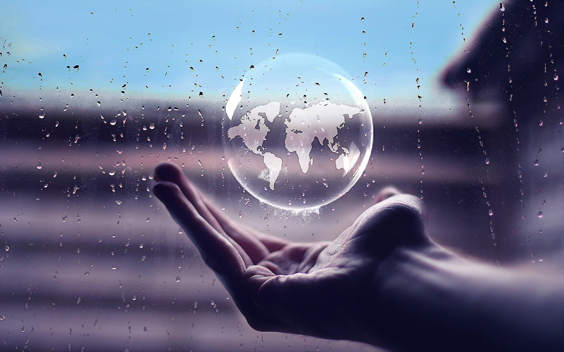 A hand holds a translucent bubble depicting a world map, set against a blurred, rainy background, blending elements of nature and artistic expression in this HD wallpaper.