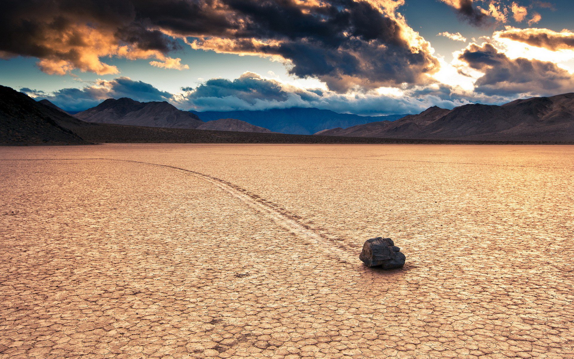 HD PC desktop wallpaper showcasing a vast desert landscape with a solitary rock leaving a trail on cracked dry earth beneath dramatic, cloud-filled skies.