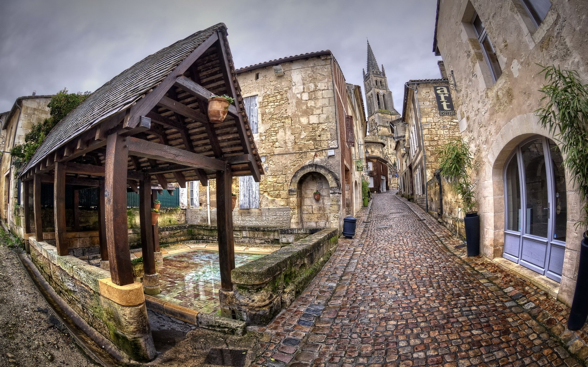 HD PC desktop wallpaper of Saint-Émilion’s man-made medieval streetscape: cobblestone lane, covered stone washhouse and distant church spire beneath a brooding sky.