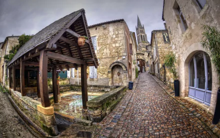 HD PC desktop wallpaper of Saint-Émilion’s man-made medieval streetscape: cobblestone lane, covered stone washhouse and distant church spire beneath a brooding sky.