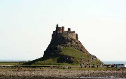 2K Quad HD PC desktop background: man-made Lindisfarne Castle perched on a rocky tidal island with grassy slopes, low tide causeway and small figures on the shoreline under a pale sky.