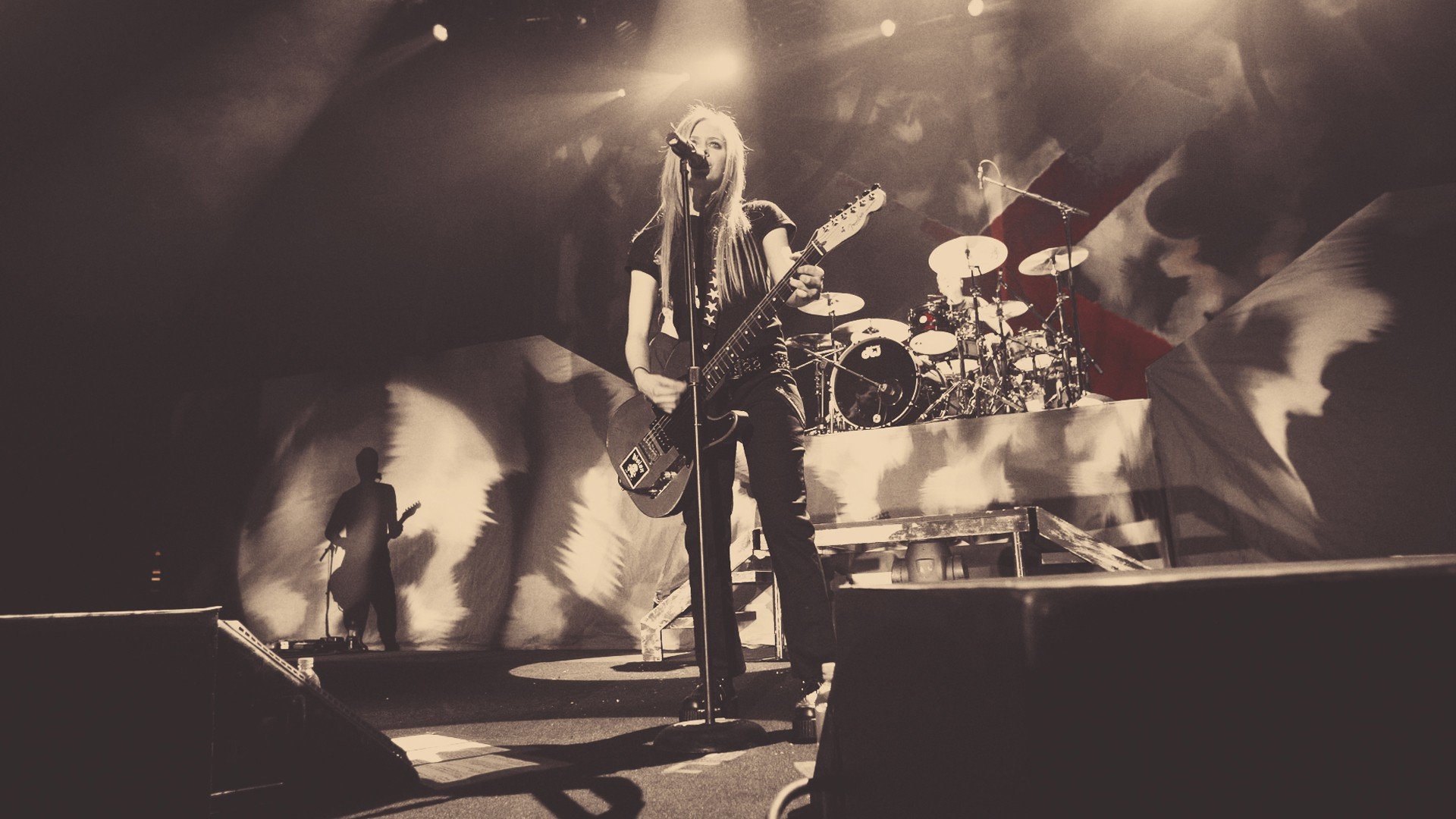 HD desktop wallpaper featuring Avril Lavigne performing live on stage, illuminated by dramatic lighting with musicians and instruments in the background.