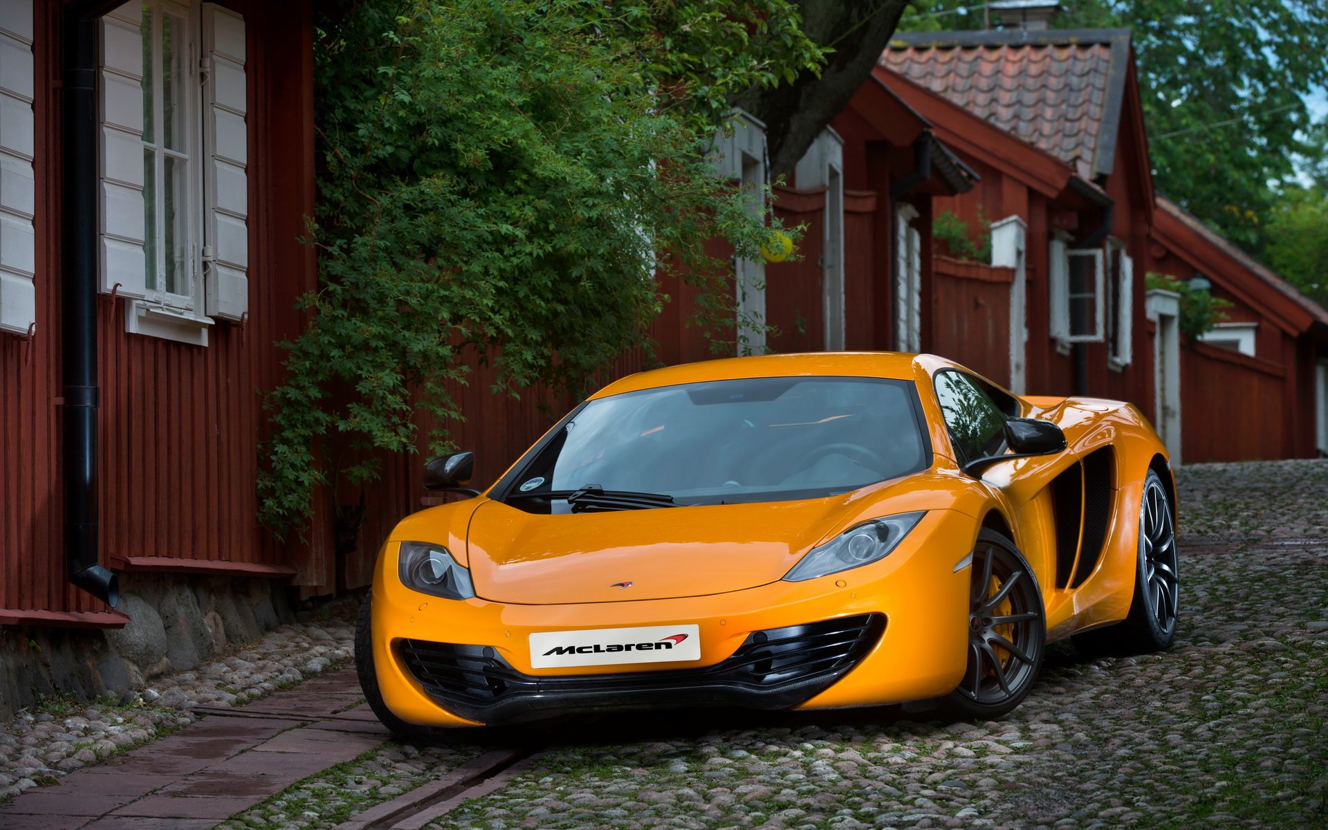 HD desktop wallpaper featuring a vibrant orange McLaren MP4-12C parked on a cobblestone street beside rustic red wooden buildings.