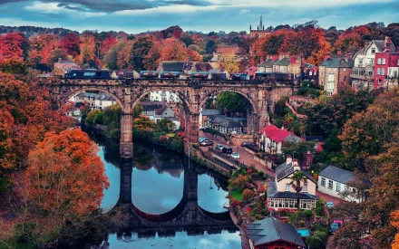 HD PC desktop wallpaper: Autumn view of Knaresborough’s man-made stone viaduct spanning a reflective river, surrounded by colorful trees and riverside buildings.