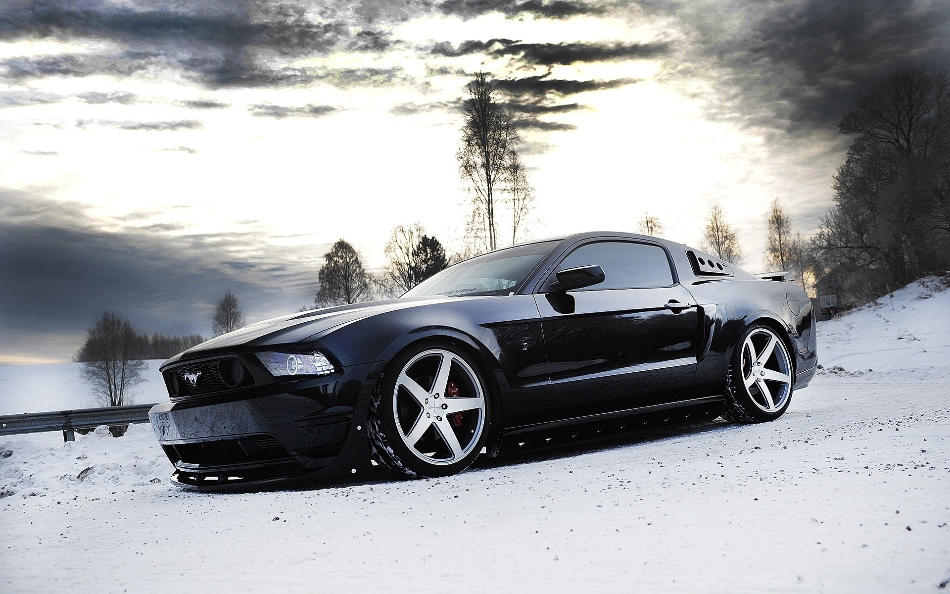 HD PC desktop wallpaper featuring a sleek black Ford Mustang parked on a snowy landscape under a dramatic cloudy sky.