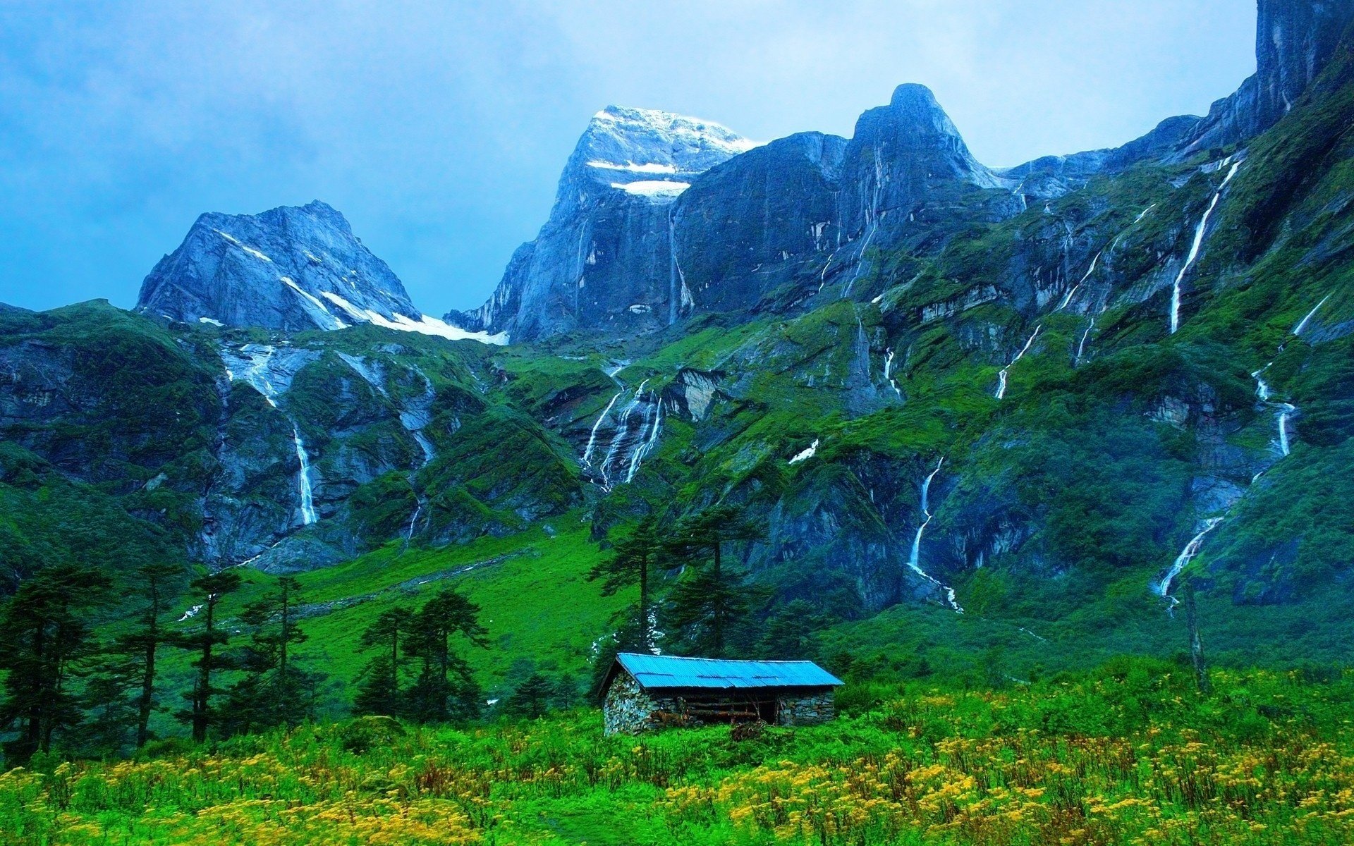 HD desktop wallpaper featuring a man-made cabin nestled in a lush green valley with cascading waterfalls and towering mountains under a clear blue sky.