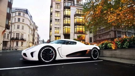 HD desktop wallpaper featuring a white Koenigsegg Agera R parked on a city street with autumn trees and buildings in the background.