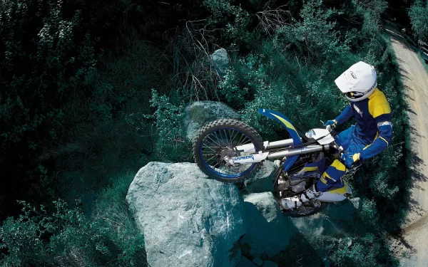 A motocross rider skillfully maneuvers over a large rock, surrounded by lush greenery, showcasing the thrill of the sport. HD desktop wallpaper for enthusiasts.