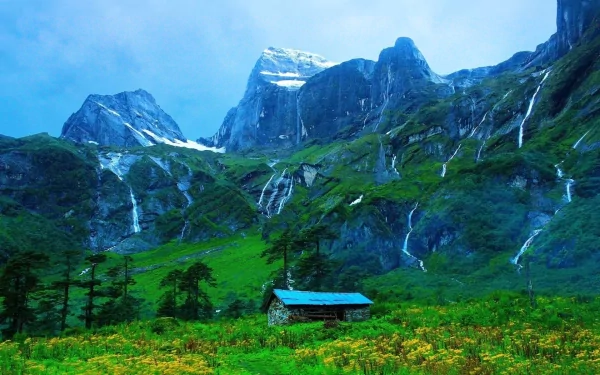 HD desktop wallpaper featuring a man-made cabin nestled in a lush green valley with cascading waterfalls and towering mountains under a clear blue sky.