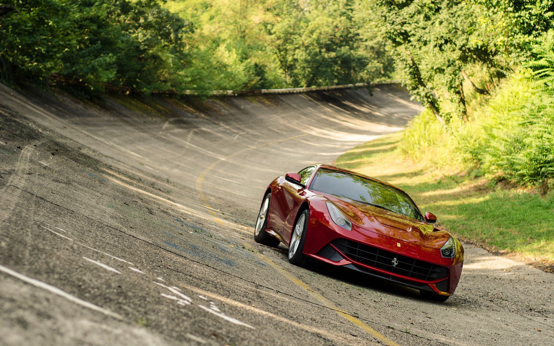 HD desktop wallpaper featuring a red Ferrari sports car driving on a winding, vintage racetrack surrounded by lush greenery.