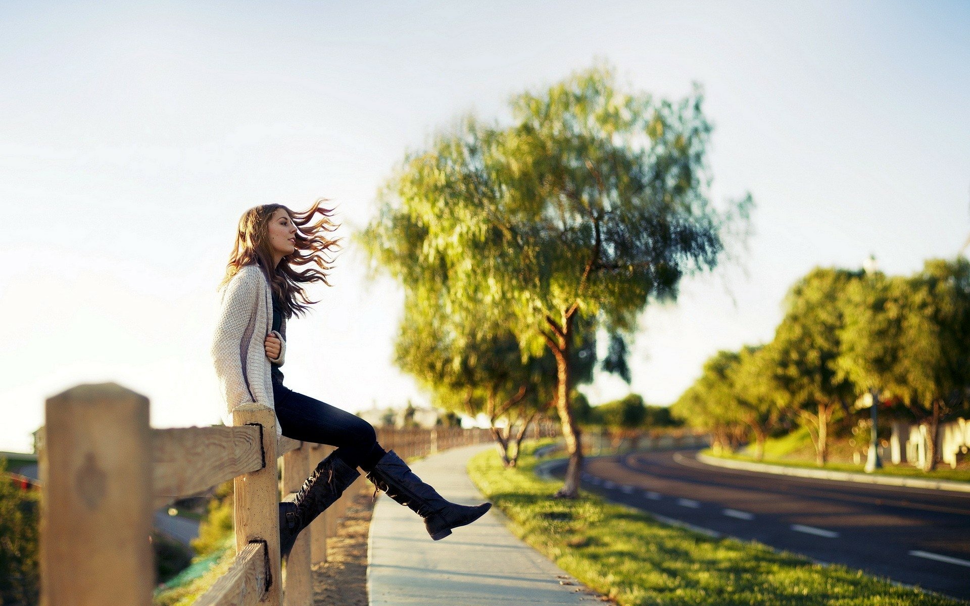 HD PC desktop wallpaper and background — woman perched on a fence by a quiet road, hair blowing, contemplative mood amid sunlit trees.