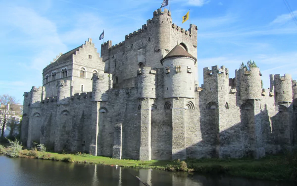 HD desktop wallpaper showing the man-made stone fortress of Gravensteen under a bright blue sky, reflected in the calm water below.