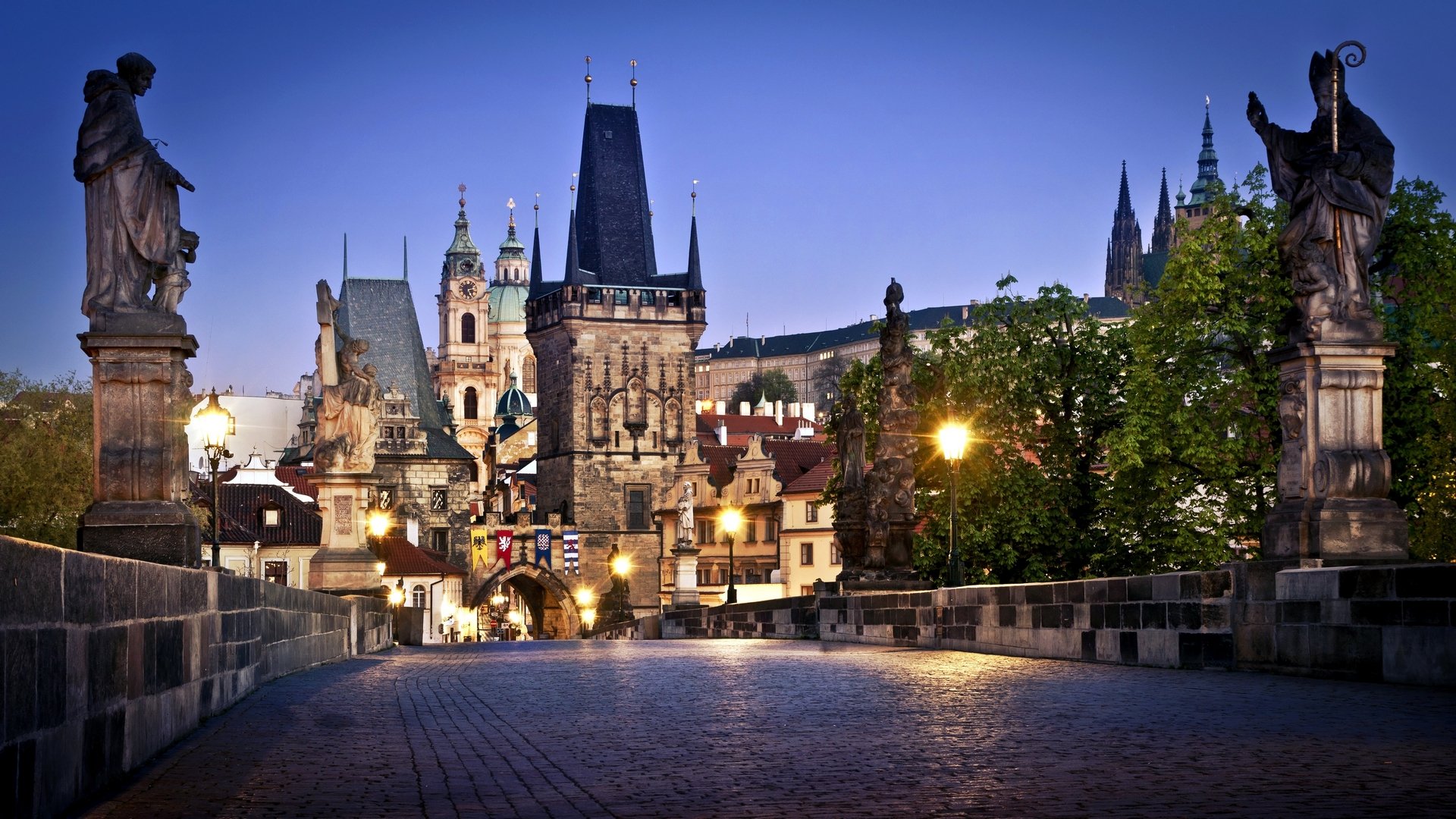 HD PC desktop wallpaper: man-made Charles Bridge in Prague at dusk, statues lining the cobblestone path with illuminated Gothic towers in the background.