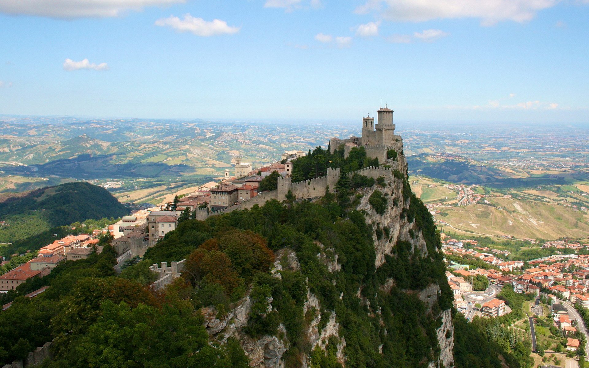 HD desktop wallpaper featuring a man-made medieval village and fortress perched atop a rocky hill overlooking a vast landscape under a clear blue sky.