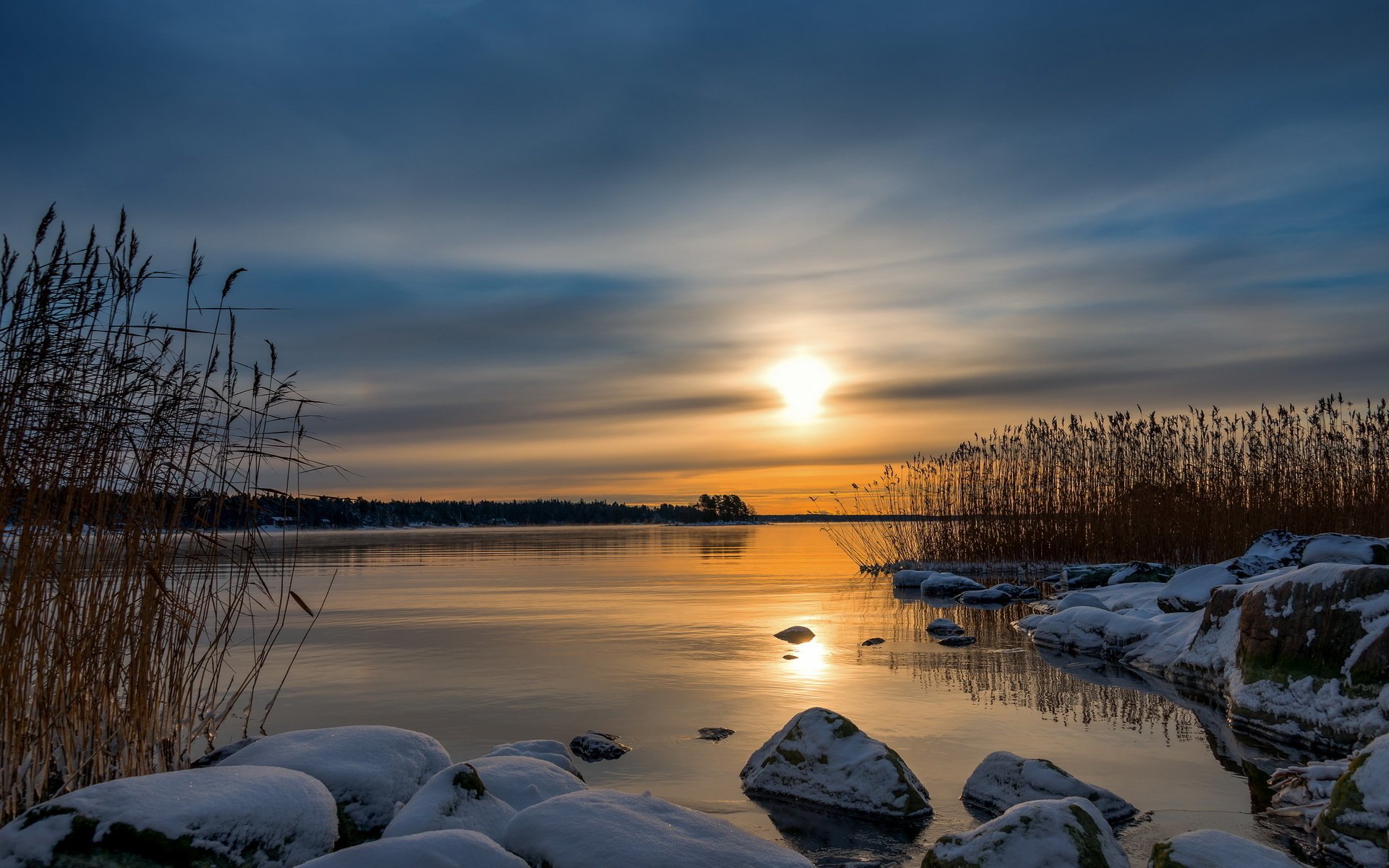 HD PC desktop wallpaper: winter lakeshore at sunset with reeds, snow-covered rocks and a golden sun reflected in calm water.