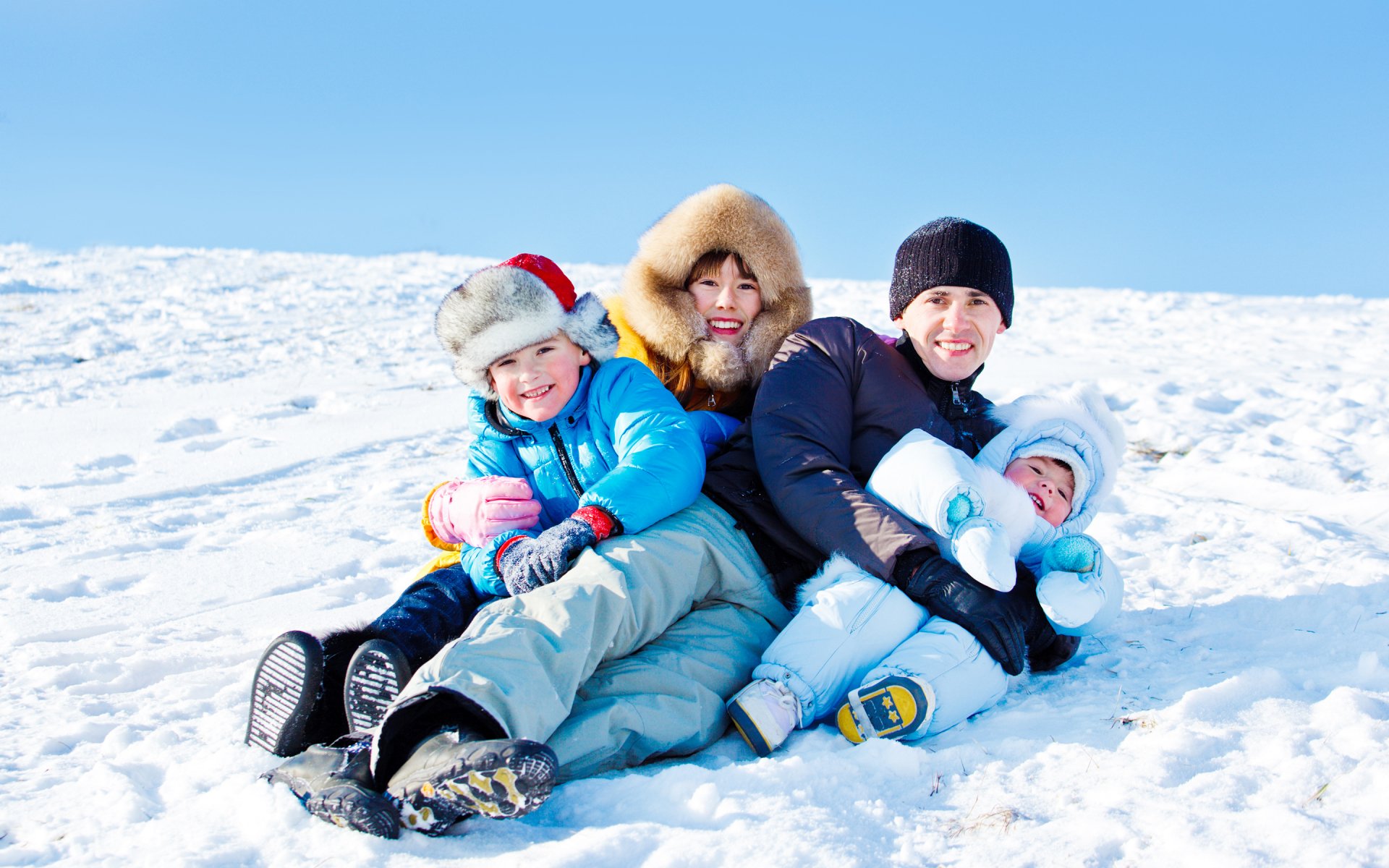 HD PC desktop wallpaper featuring a joyful family of four dressed in winter clothes, sitting and playing together on snow under a clear blue sky.
