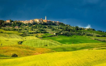 HD desktop wallpaper of Pienza, a man-made hilltop town in Tuscany, Italy, surrounded by vibrant green and yellow fields under a clear blue sky.
