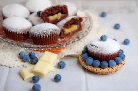 A beautifully arranged display of cupcakes, some filled with cream and dusted with powdered sugar, surrounded by fresh blueberries and white chocolate, serving as a vibrant HD desktop wallpaper.