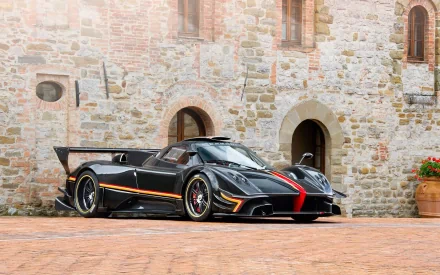 HD PC desktop wallpaper featuring a sleek black Pagani Zonda R sports car parked on a brick driveway in front of a rustic stone building.